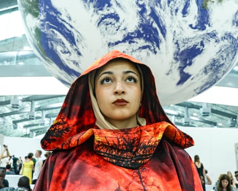 A woman in an orange hooded cape showing flames and burnt trees stands beneath a giant globe in a conference centre