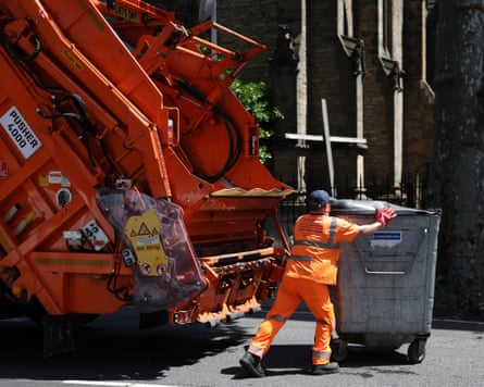 A refuse worker in hi-vis orange trousers and jacket positions a bin behind a bin lorry