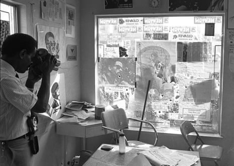 A man takes a photograph of a window with a bullet hole in it, which is also covered in newspaper and other printed material