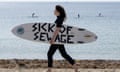 A woman carrying a surfboard that reads 'Sick of sewage' walks along a beach as people surf in the background