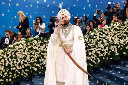 A man wearing a white turban, cloak and trouser suit with gold and emerald accessories and a ceremonial sword, stands on a flight of blue-carpeted stairs in front of a press pack.