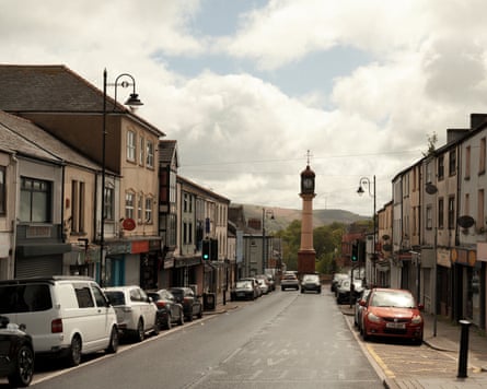 A think painted iron clock tower at the end of a row of shops and houses, a hillside behind