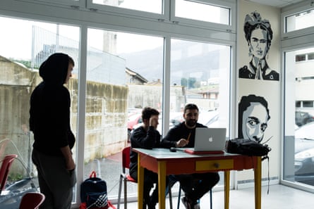 A teenagers sits at a desk next to a man with a laptop on the table as another teenager approaches