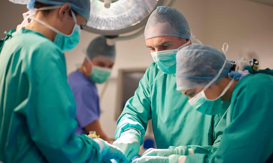 A female surgeon, dressed in scrubs, stands over a patient at the operating table, looking down at what she is doing in the operation. She is joined by a female nurse and male surgeon, while in the background, an anaesthetist looks over from his monitors . They are all wearing surgical gowns and protective masks and headwear.
