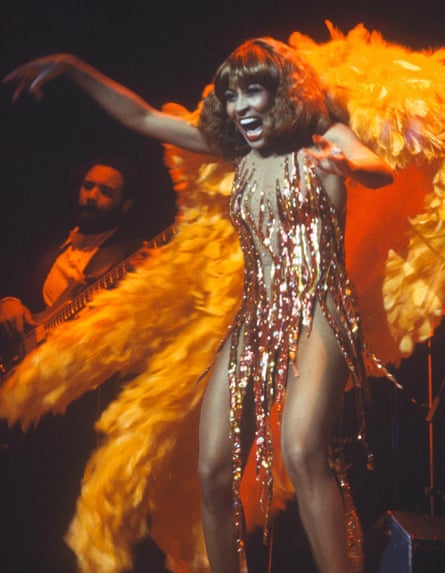 A woman wearing a sparkly illusion dress with orange feathers to the back singing and dancing
