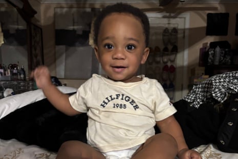 Kohen Wiley, a smiling baby in a cream t-shirt, sits on a bed waving