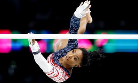 A sideways picture of Simone Biles, in a stars and stripes leotard and wearing partial supports on her palms, leaps towards a pole