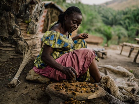 A woman sits on the ground and grinds nuts on a piece of wood.