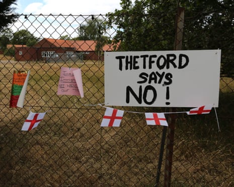 A sign attached to a wire fence reads Thetford Says No! There is a string of small England flags underneath it, and other messages pinned to the wire. There are low-rise brick buildings behind the fence.