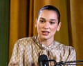 Dua Lipa speaks into a podcast microphone while wearing a patterned beige shirt in front of a gold curtain