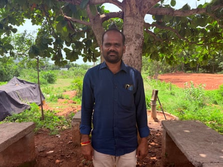 A man stands in front of a tree, with simple benches either side of him.