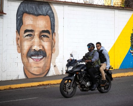 Two men on a motorcycle pass a mural of Nicolás Maduro