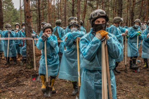 People in helmet, goggles, face mask and blue plastic overalls rest on shovels in a wooded area