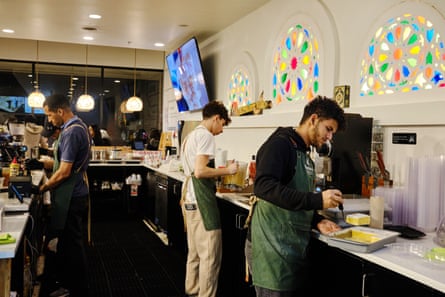 three barista behind a counter making drinks and cutting cake, beneath colourful stained-glass windows