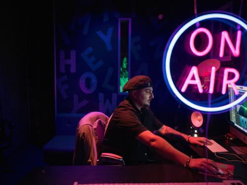 Man wearing a cap backwards in a dark room looking at a computer screen, with a pink neon ‘on air’ sign in the foreground.