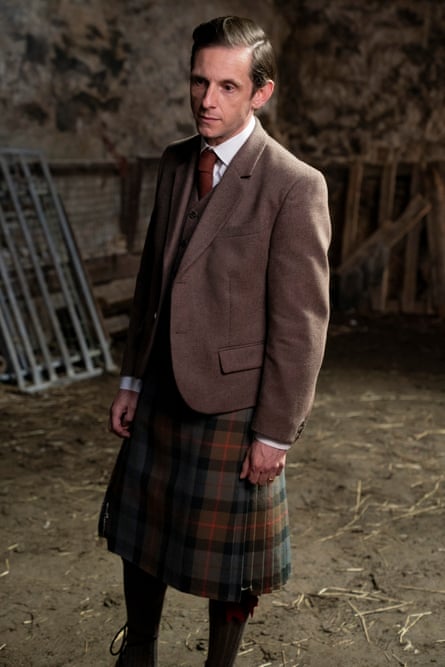 A man in a brown kilt and formal jacket standing in a barn