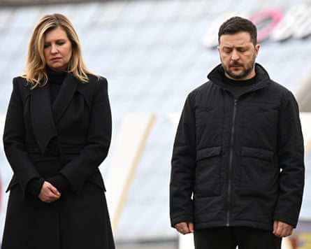 Zelenska and Zelenskyy stand solemnly with their heads bowed