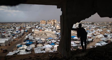 A person is silhouetted standing inside a destroyed building overlooking a makeshift camp with damaged buildings in the background