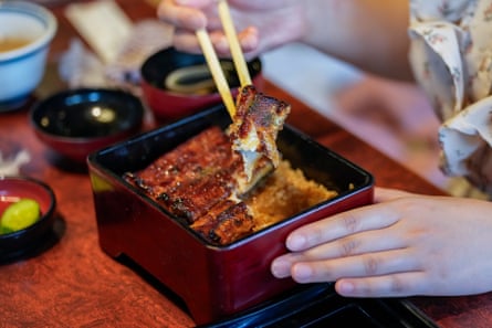 A square bowl of Kabayaki-style eel on a restaurant table with several small round dipping bowls next to it. A person’s hand can be seen lifting a piece of fish out of the bowl with chopsticks