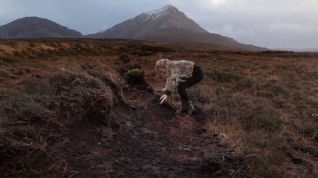 A woman in a fur-textured jacket crouches, reaching toward muddy ground, with a mountain behind her