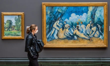 A woman wearing a face mask looks at the a painting of naked people sitting on the ground against a blue background