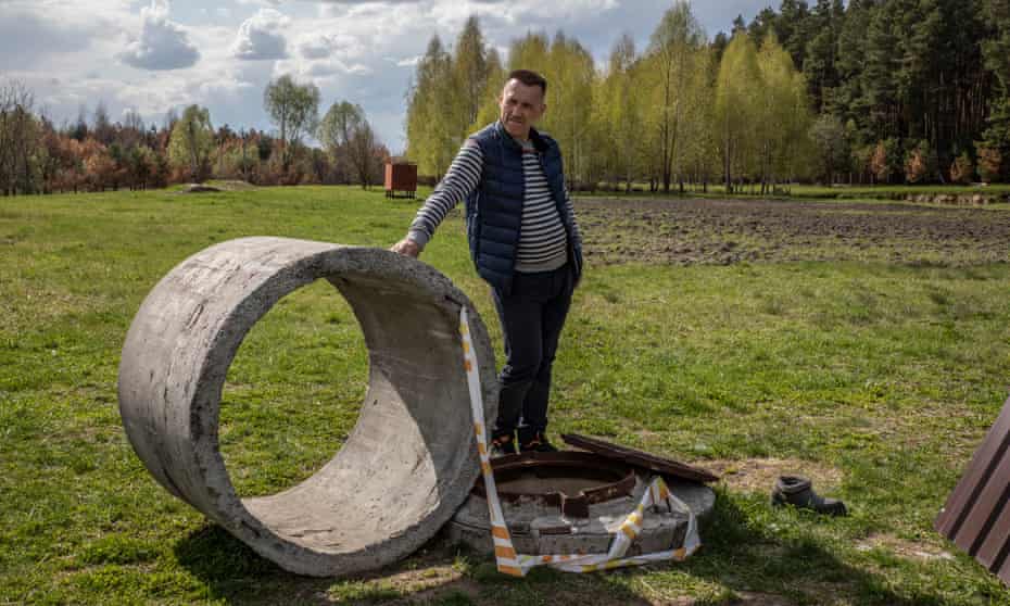 A man, looking off camera,  leans on a concrete construction cylinder by a small uncoverd manhole sunk into a field, with trees in the background