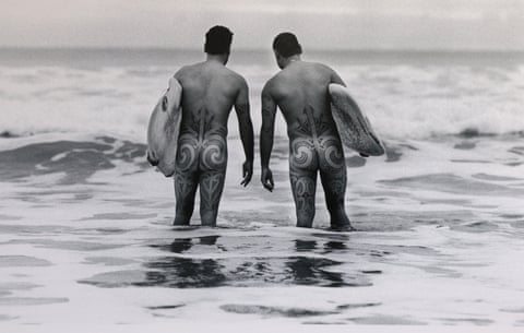 Two naked men with intricately tattooed legs and buttocks carry surfboards in knee-deep water
