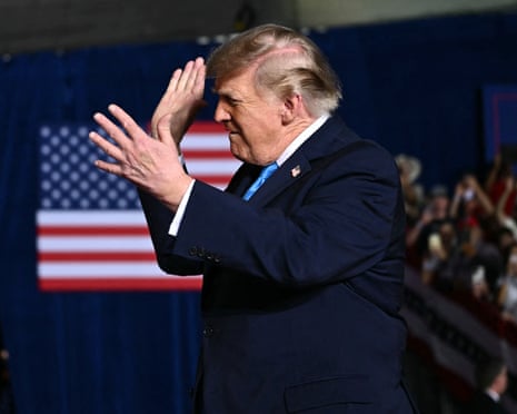 Trump gesturing with his hands, in front of a crowd and a US flag