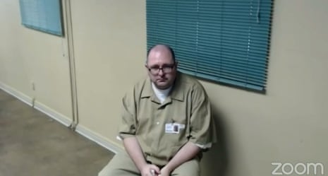 A man wearing khaki pants and shirt sits in an empty room in this screen grab from a Zoom video call.