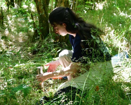A woman kneels down in the dappled light of a forest holding a silver-coloured cylinder.