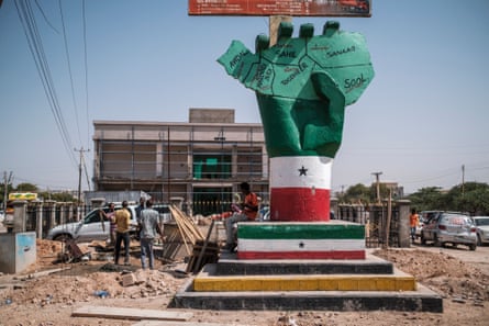 People stand next to a sculpture of a hand holding a map of the country, located by the side of a busy road surrounded by building work and rubble.