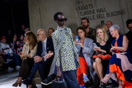 A model in a patterned green coat and sunglasses walks past onlookers and a sign that reads: ‘Tickets. Turbine Hall. Natalie Bell building’