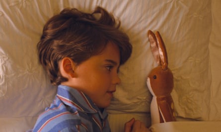 a young boy lying in bed in pyjamas looking at a velveteen rabbit on the pillow next to him