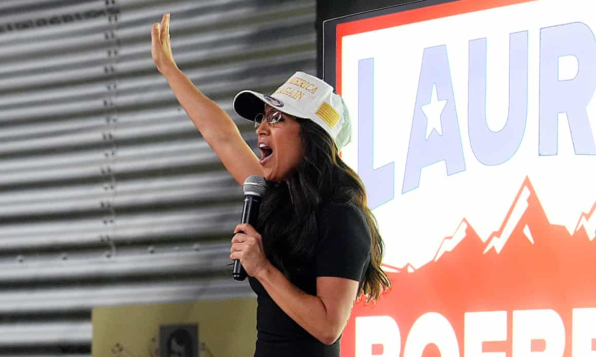 Lauren Boebert, wearing a cap, holds a microphone in one hand and holds the other in the air at a primary election watch party.