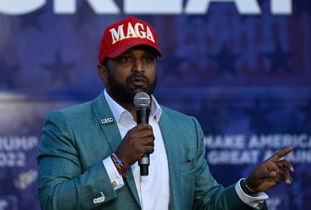 A man in a tight-fitting suit with a Maga hat on speaks into a microphone.