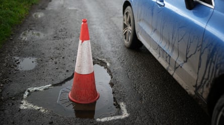 A red and white traffic cone is placed in a large, deep and water-filled pothole on a dark grey tarmac road, with white paint indicating the hole’s edges; the door, wings and rear wheel of a dark blue car are seen passing the hole; the side of the car is streaked with water and mud and dirt