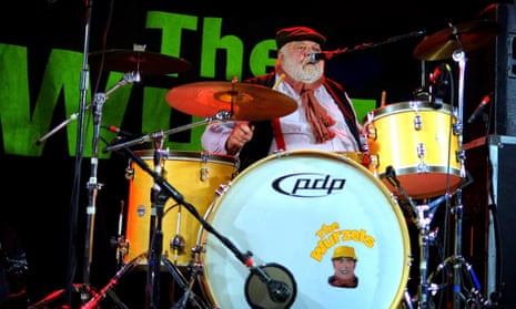 John Morgan on stage behind a yellow drumkit wearing a cloth cap and scarf