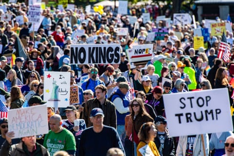 wide shot of a large group of people at a rally holding signs that include 'make America democratic again' 'stop Trump' and 'no kings'