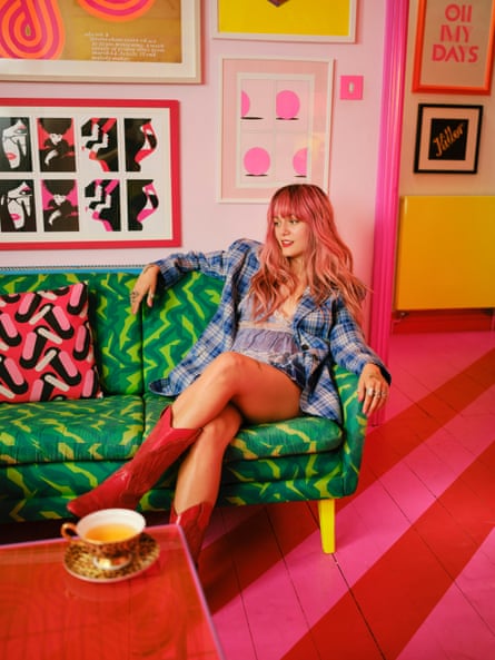 Florence Given sitting on her green patterned sofa dressed in cowboy boots, shorts, and a demin jacket. The floor in pink and red and the walls are covered in artwork