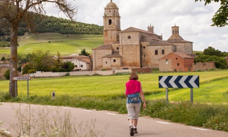 A female pilgrim walking on a narrow country road approaches a historic church on the edge of a town on a green hillside