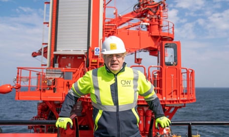 Man looking uncomfortable in hard hat and hi vis clothing, in front of some bright orange machinery and a calm sea
