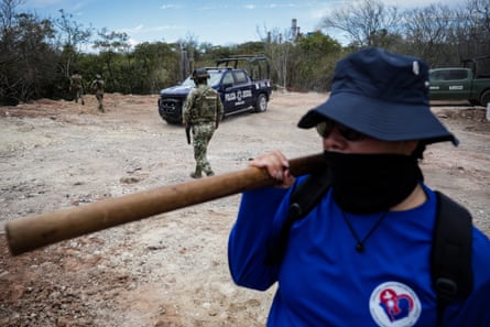 A person in hat and mask carries a pickaxe while a uniformed agent walks in the background near a car.