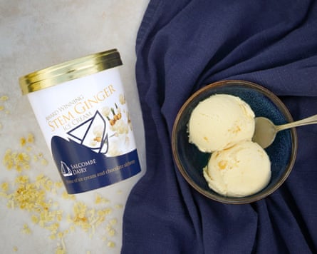 A tub of Salcombe Dairy Stem ginger ice-cream Press publicity image on its side next to a blue cloth with a bowl containing two scoops of ice-cream and a spoon