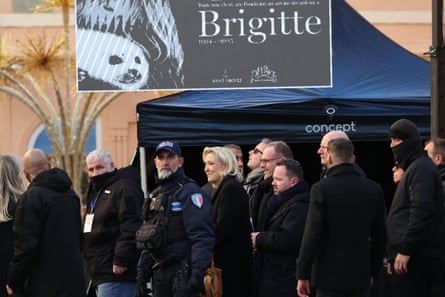 Marine Le Pen, dressed in black, stands in a group of people and behind a police officer; on the wall behind her there is a large black and white poster showing Bardot holding a seal.