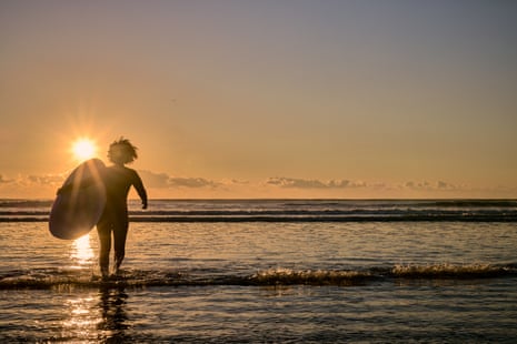 A woman surfer wearing a wetsuit and carrying a surfboard, wading into ocean water during sunset