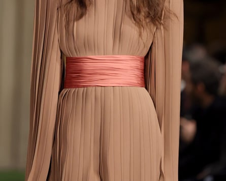 A model walks the Valentino runway wearing a pleated tan tunic with a wrapped pink belt
