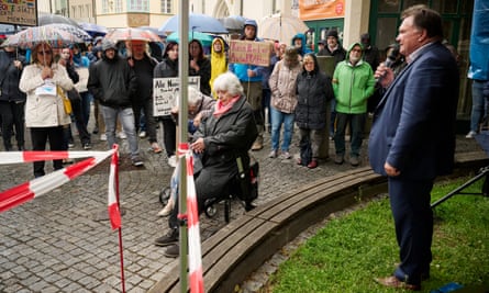 Stefan Bosse, in a suit, stands on a patch of grass holding a microphone and addressing a seated older woman and members of the anti-AfD rally holding umbrellas