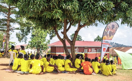 A group of people wearing matching yellow tops sit under a tree outside a building, next to a branded event flag