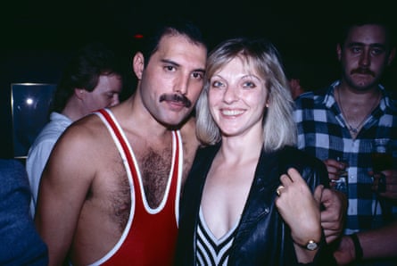 A man with dark hair and a moustache wearing a red singlet with his arm round a blond woman wearing a black jacket and black and white top