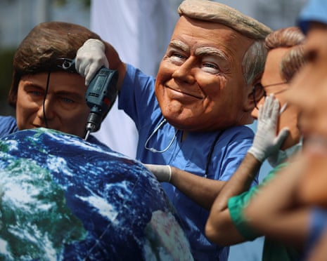 people wear oversized masks depicting world leaders, including one holding a power drill over a ball representing the earth